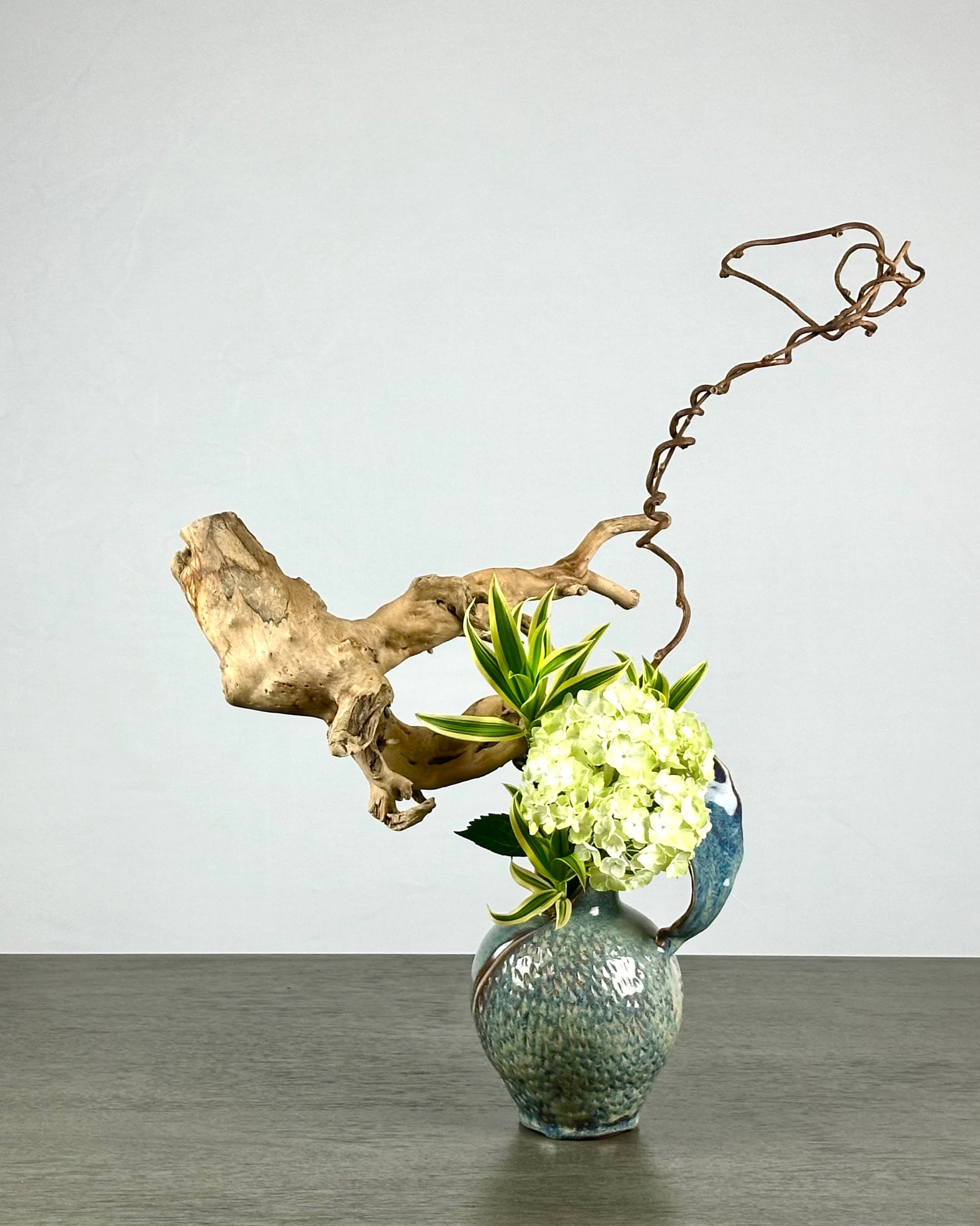 Decorative arrangement with a ceramic vase, greenery, white hydrangea, and driftwood on a gray surface.